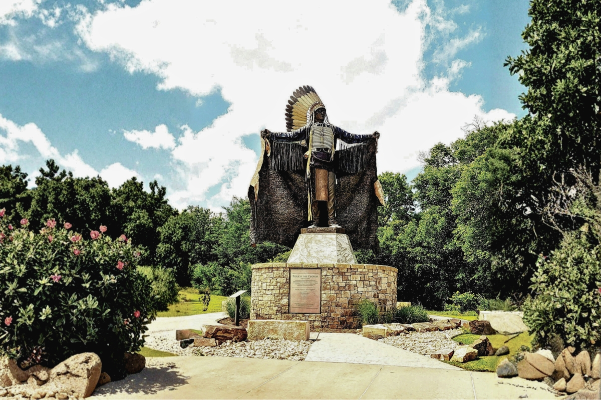 Statue of a Native American figure with outstretched arms in a landscaped park setting in Edmond, Oklahoma