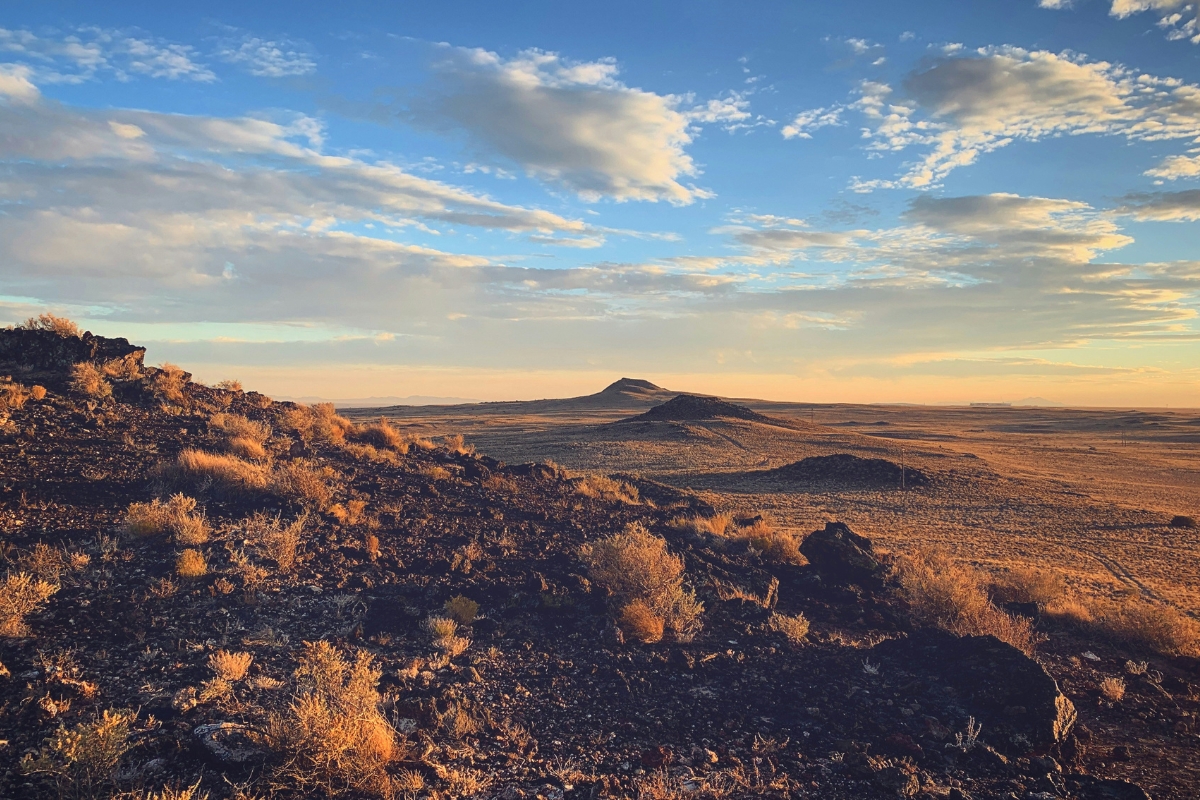 Rocky desert landscape in New Mexico with scrub vegetation and low mesas under a partly cloudy sky at sunset