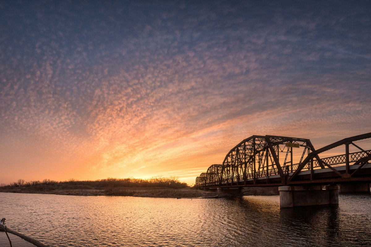 Steel truss bridge over a river in Oklahoma at sunset with dramatic cloud patterns and warm reflected light on the water