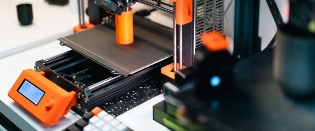Desktop 3D printer fabricating an orange plastic cylindrical prototype on a build plate in a workshop setting