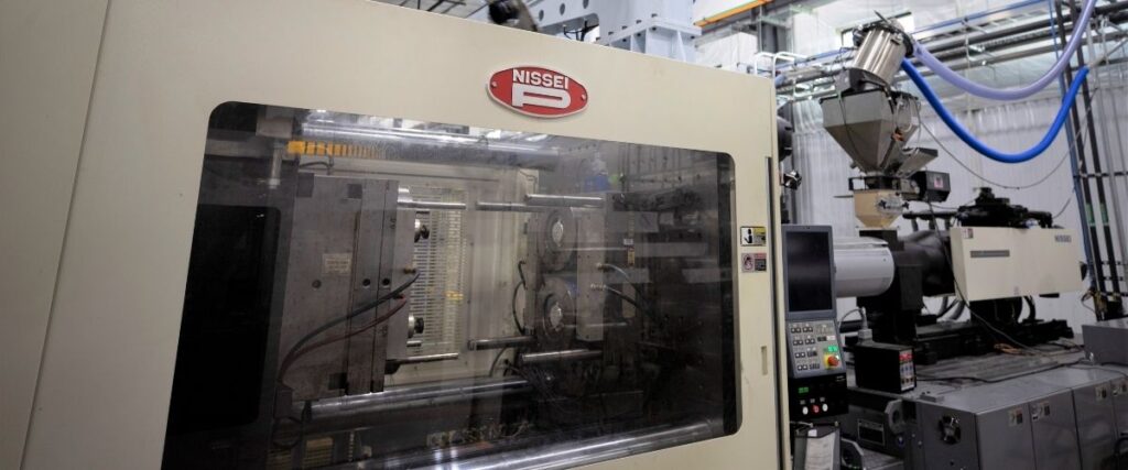 Injection molding machine operating in a manufacturing facility with mold tooling visible inside the press and material feed system attached