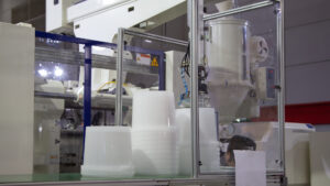 Automated injection molding press producing stacked white plastic containers in a manufacturing facility.