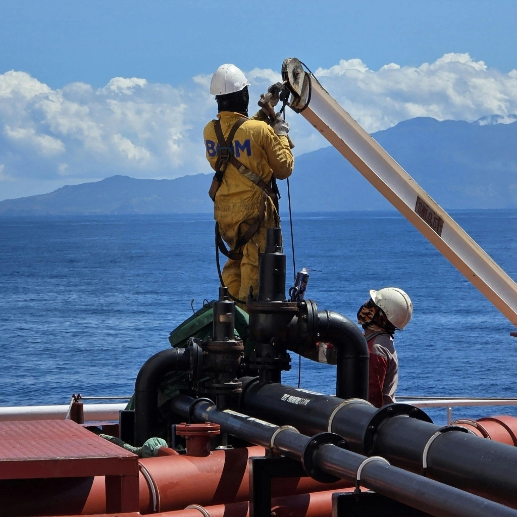 Workers wearing hard hats perform maintenance on offshore pipeline and valve equipment with ocean and mountains in the background.