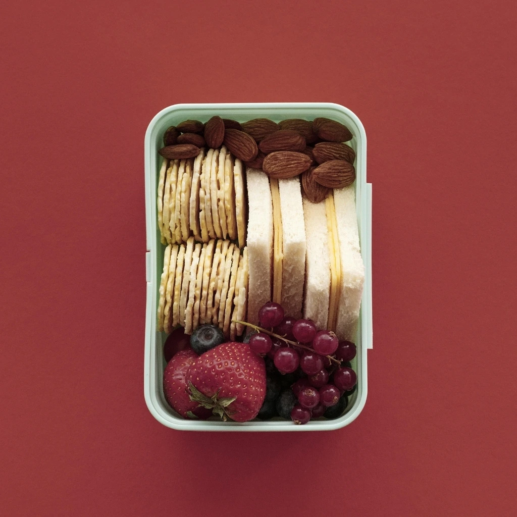 Reusable plastic food container filled with crackers, sandwich slices, almonds, strawberries, blueberries, and grapes.