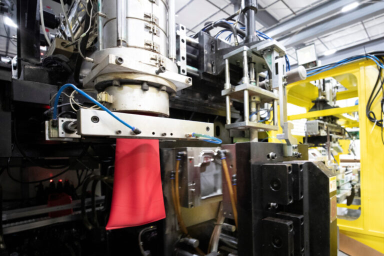 Injection molding machine producing a red plastic component inside a manufacturing facility with tooling, automation, and safety guarding visible.