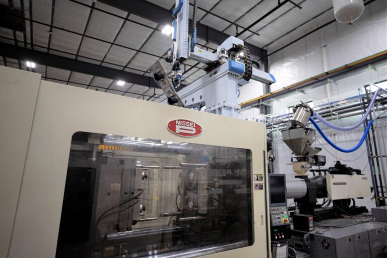 Automated Nissei injection molding machine with robotic part handling, material feed equipment, and enclosed tooling inside a plastics manufacturing facility.