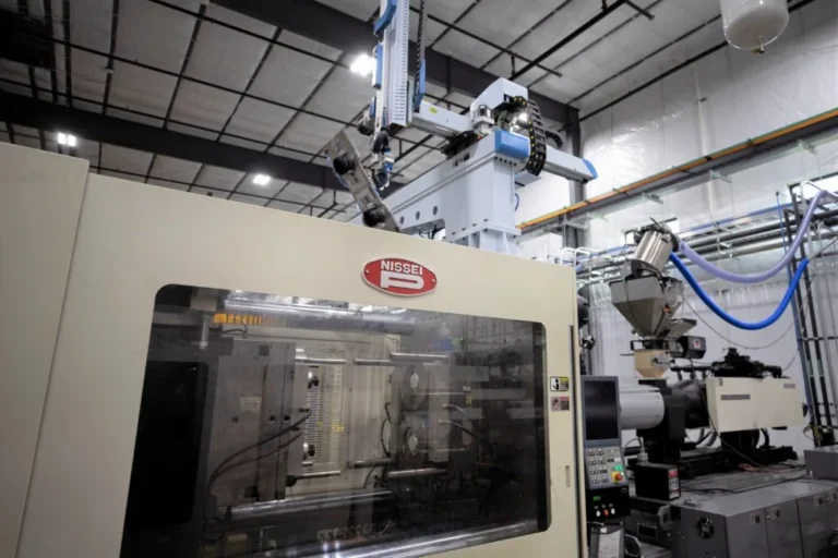 Automated Nissei injection molding machine with robotic part handling, material feed equipment, and enclosed tooling inside a plastics manufacturing facility.