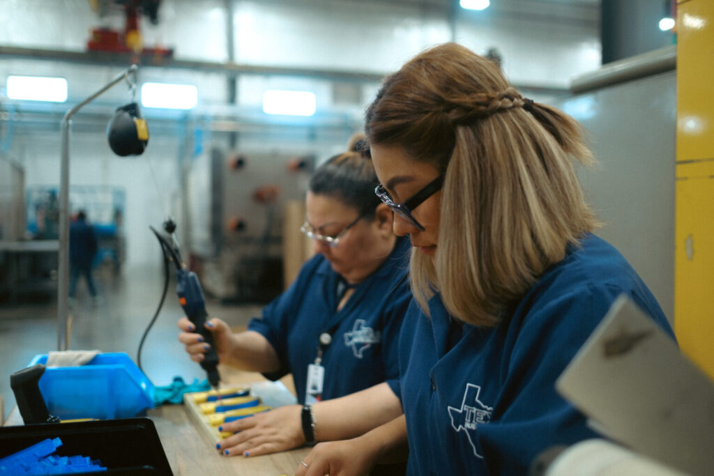 Manufacturing team members wearing safety glasses inspect and assemble small plastic components at a workstation.