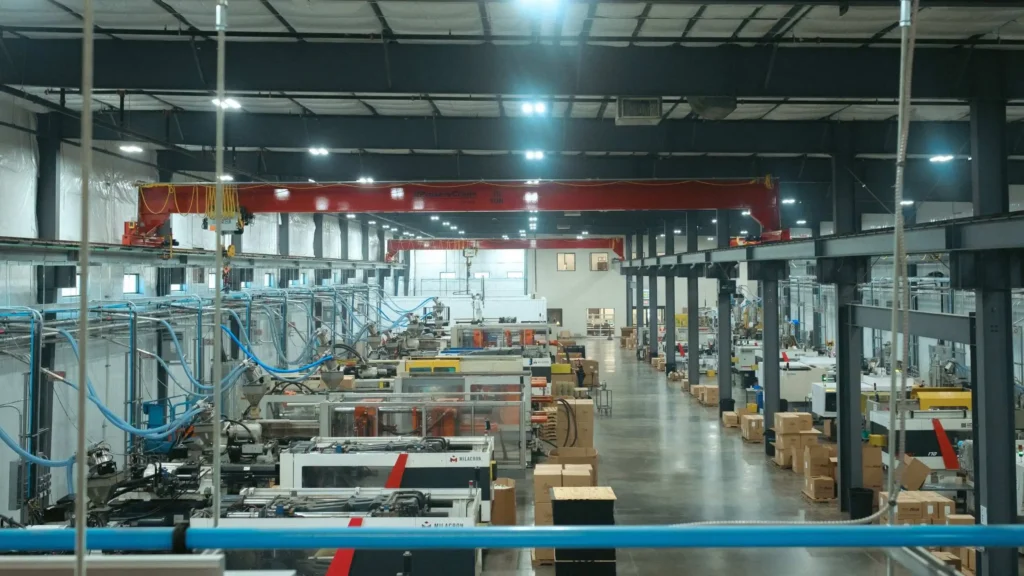 Wide view of an injection molding facility with multiple molding machines, overhead crane, material lines, and boxed parts