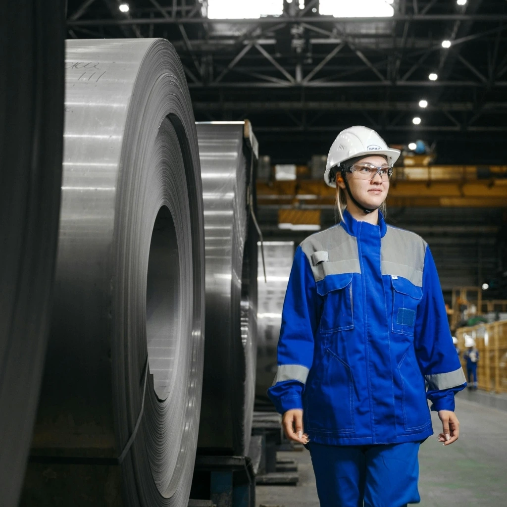 Manufacturing worker wearing a hard hat and safety glasses stands beside large steel coils inside an industrial production facility.